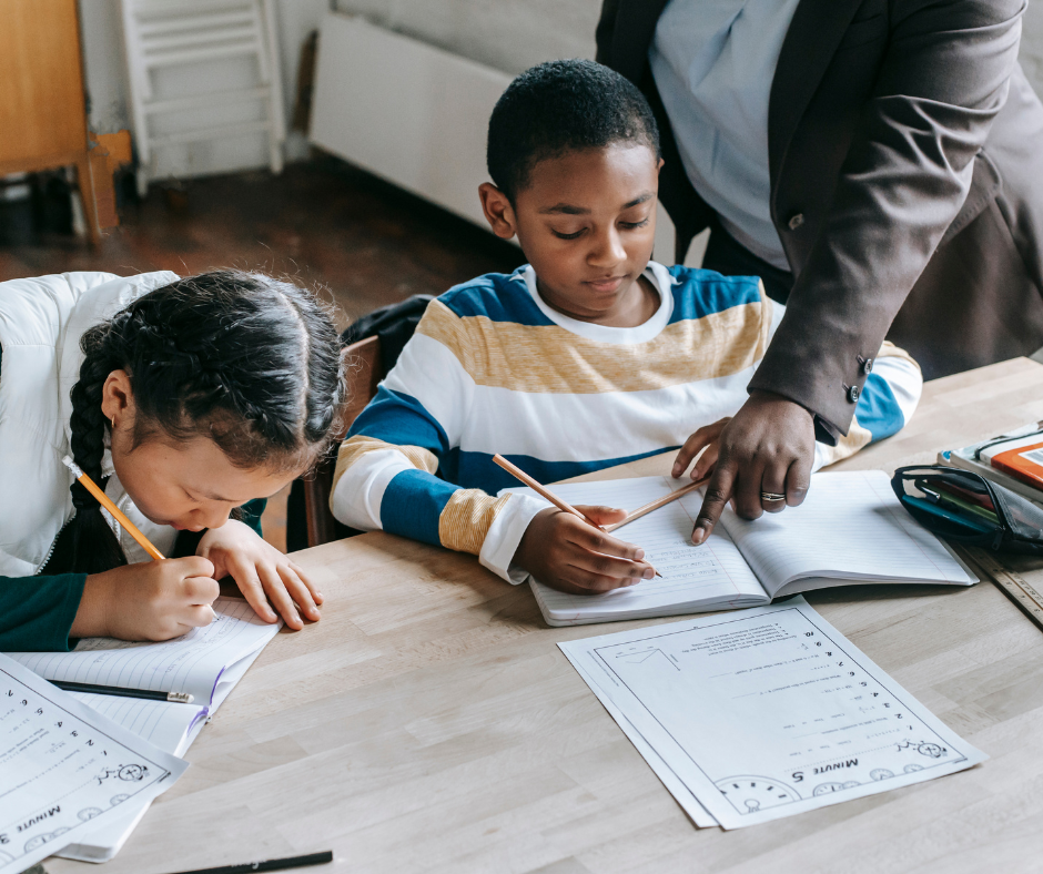 Two children writing at a desk with guidance from an adult.