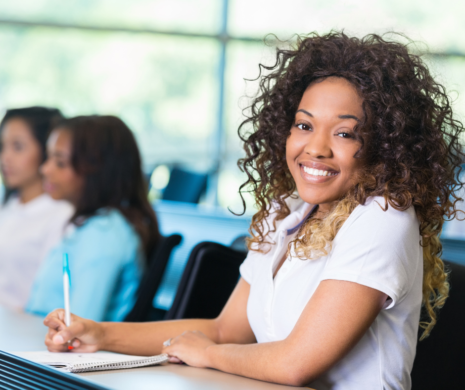 Smiling young woman sitting in a classroom, taking notes with classmates in the background.