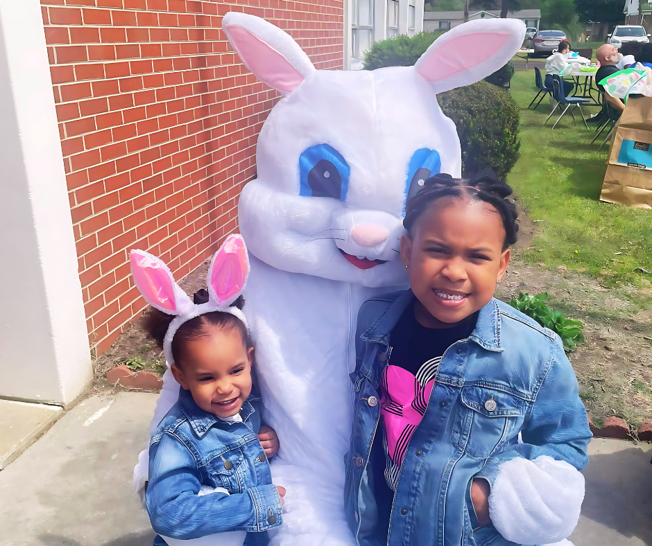 Two smiling children wearing bunny ears posing with a person in an Easter Bunny costume outdoors.