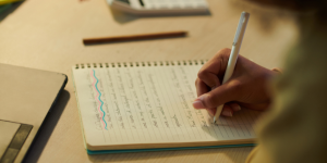 Close-up of a person writing in a notebook with a laptop on the desk.