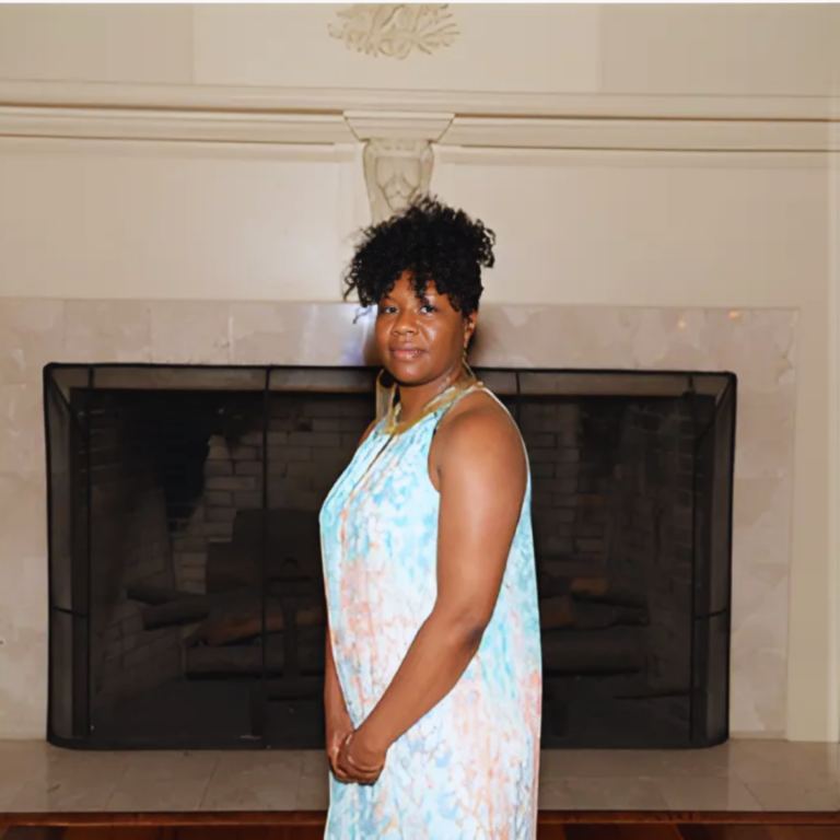 Ann Slater standing in front of a fireplace, wearing a sleeveless light blue dress with a floral pattern.