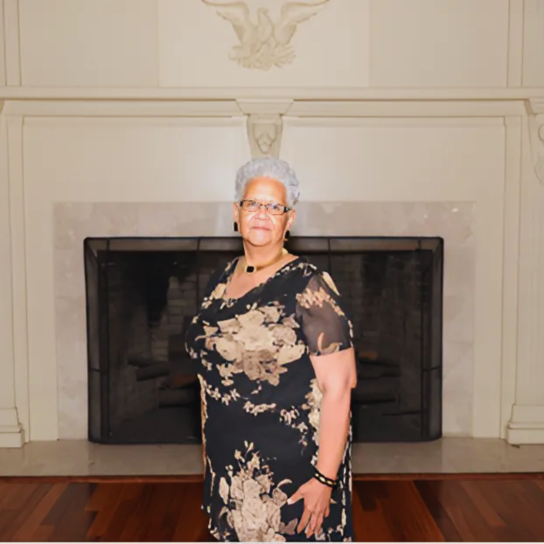 Barbara Ridley smiling in a floral dress, standing in front of a fireplace.