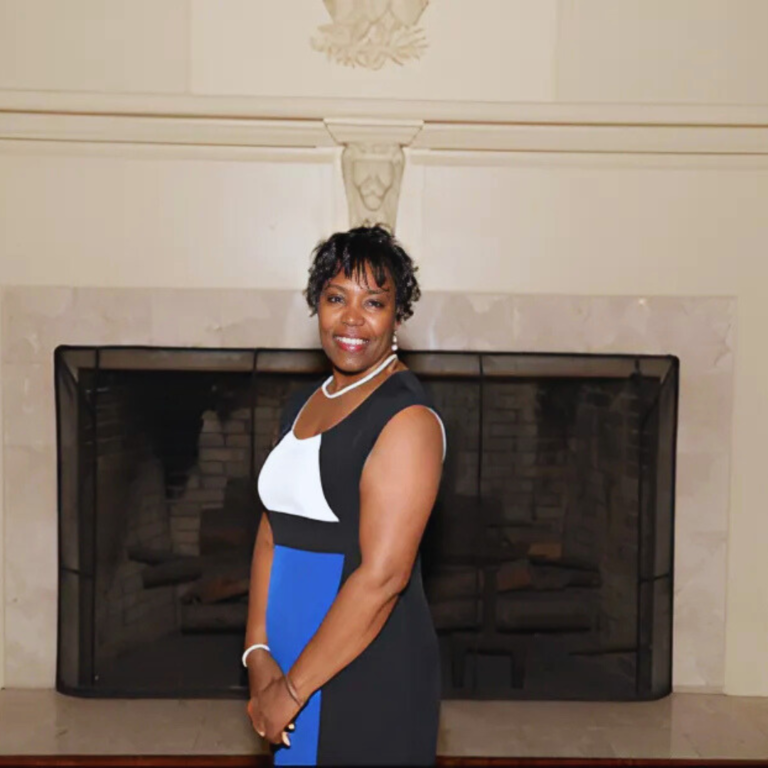 Dr. Phyllis Moore Tolliver smiling and standing in front of a fireplace, wearing a sleeveless black, white, and blue dress.
